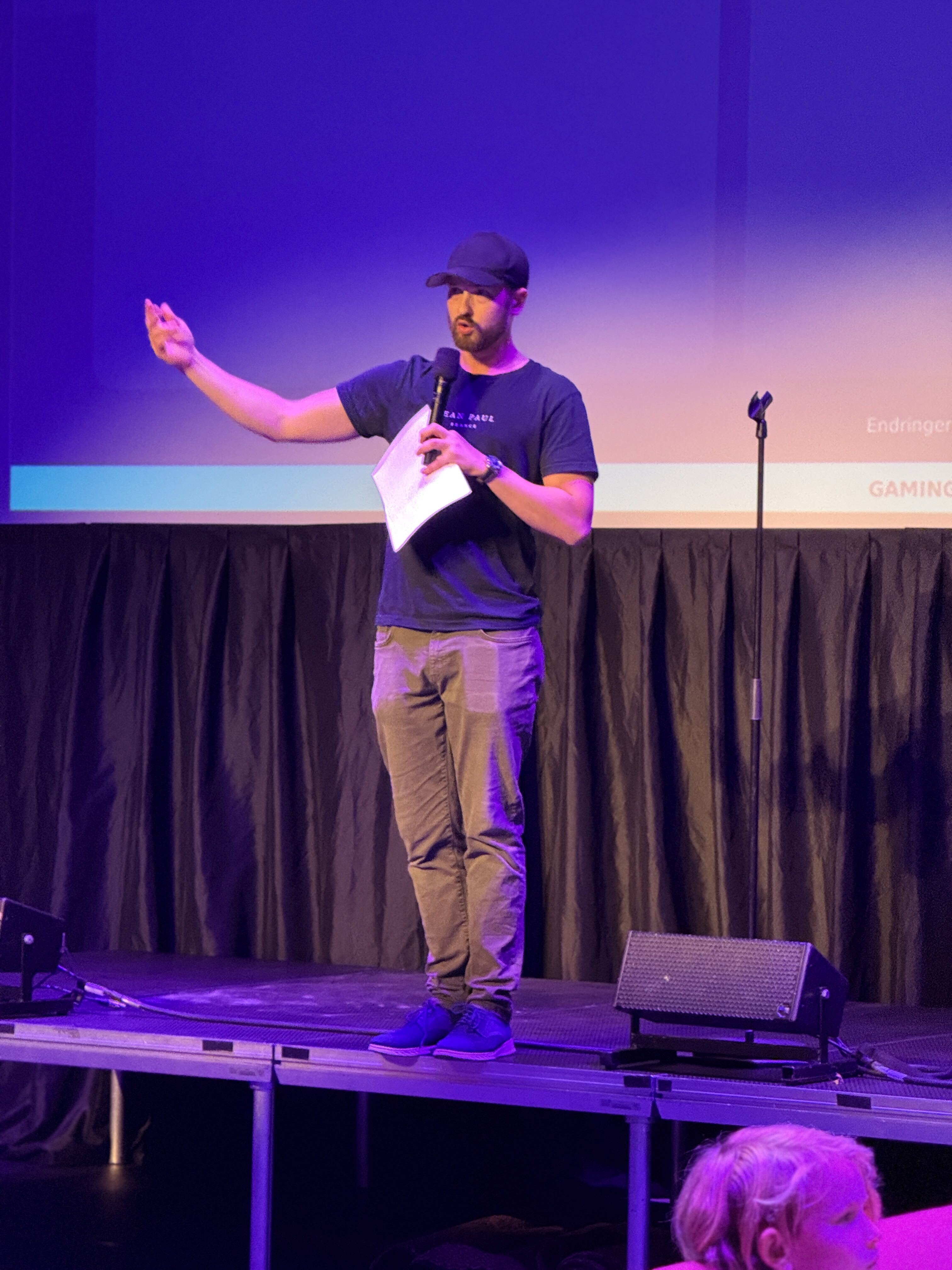 Fredrik Grøslie med mikrofon på en scene belyst med lilla lys under LEVEL UP LAN Kongsberg