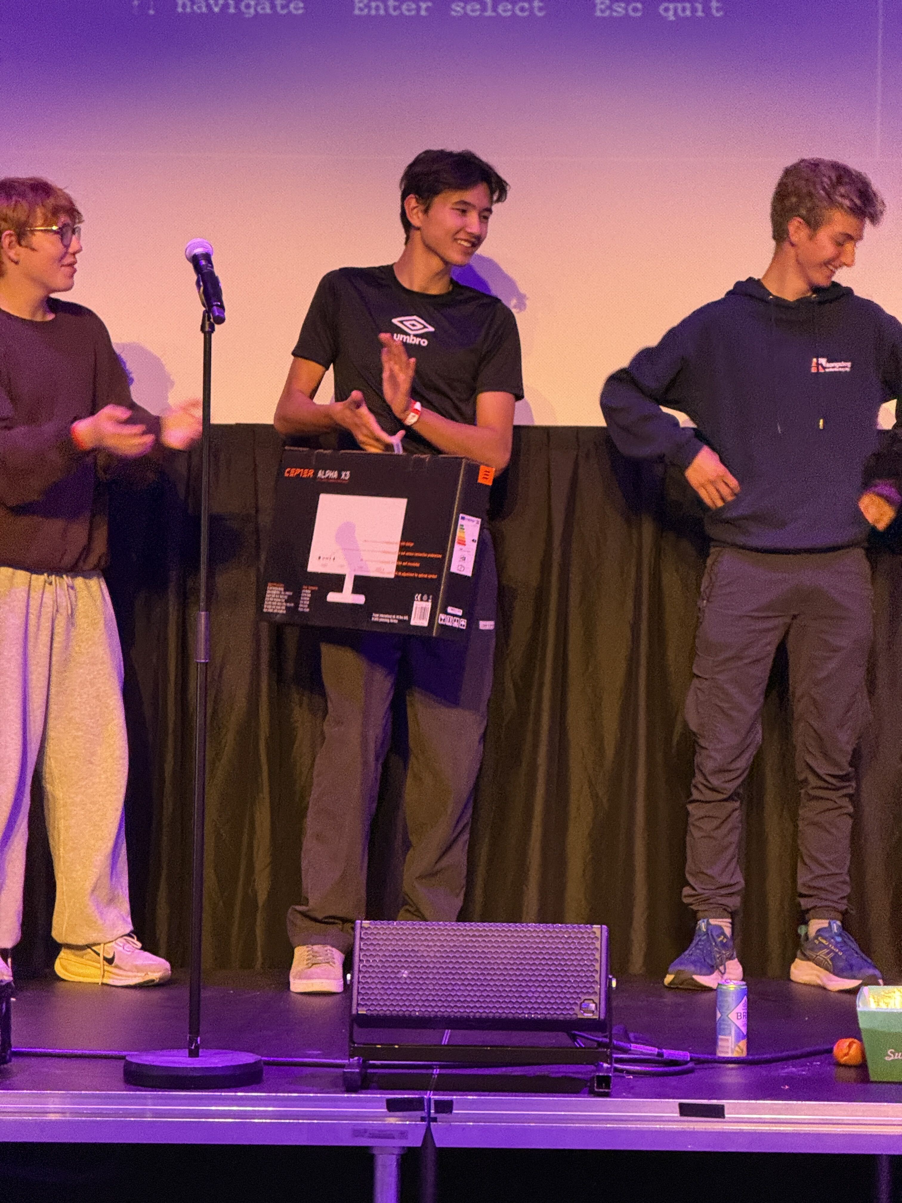 Tre personer på scenen under LEVEL UP LAN Kongsberg, der én holder en eske med bilde av en skjerm